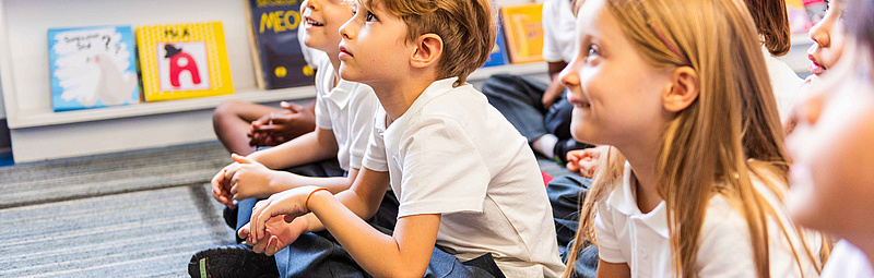 Eine Gruppe von Kindern sitzt am Boden und hören jemandem zu, der einen Vortrag hält