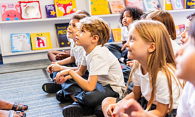 Eine Gruppe von Kindern sitzt am Boden und hören jemandem zu, der einen Vortrag hält